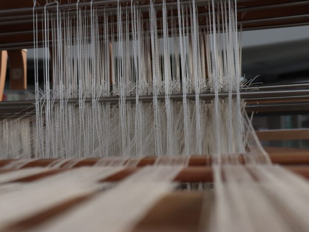 white threads being weaved into fabric on industrial loom.